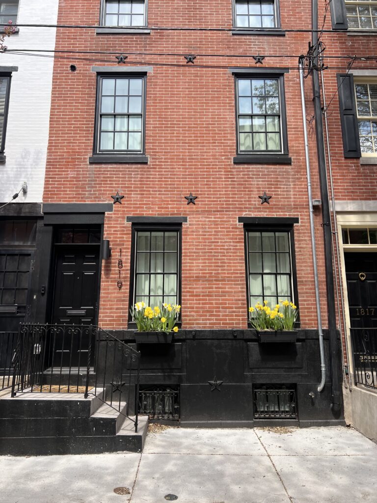 A Philly rowhome with daffofil flowerboxes and a brick facade with black accents including a black door, stoop, windows and matching black star bolts. Image by Cara Stapleton for Solo Real Estate.