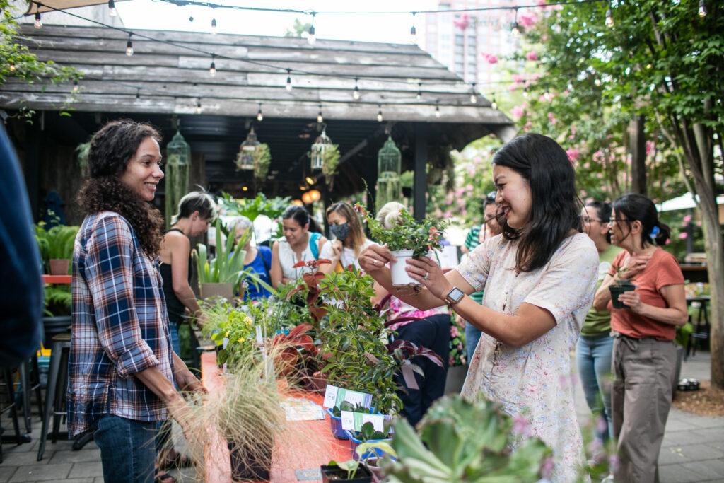 Woman holds up plant while fellow plant enthusiasts meet up for a monthly Plant Swap at the PHS Pop Up Garden courtesy of Solo Real Estate.